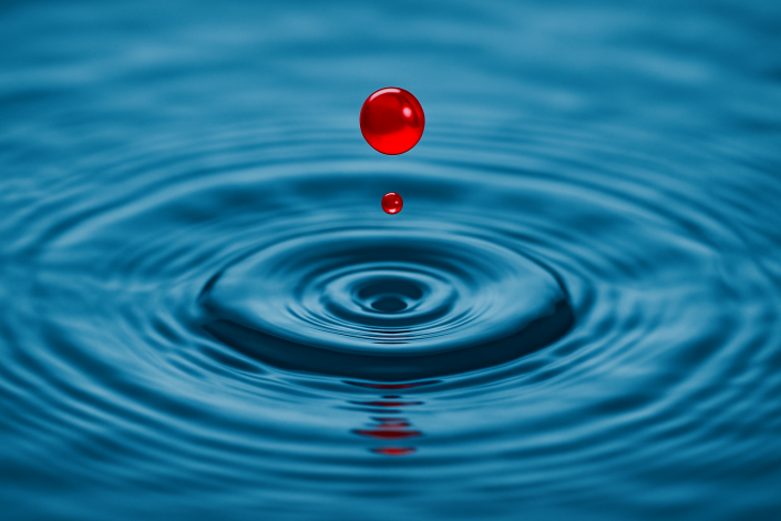 Ein roter Wassertropfen fällt ins Wasser und löst konzentrische Wellen aus.