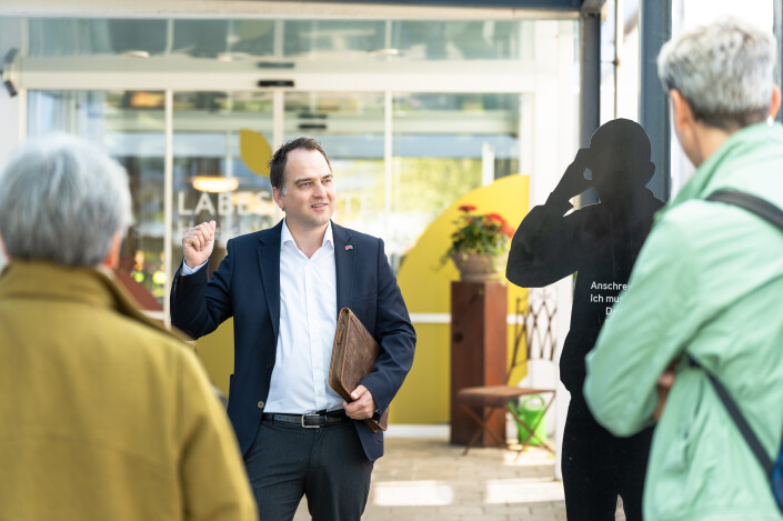 Stefan Beyeler im Anzug spricht vor einer Gruppe, im Hintergrund Eingang mit «Läbesgarte»